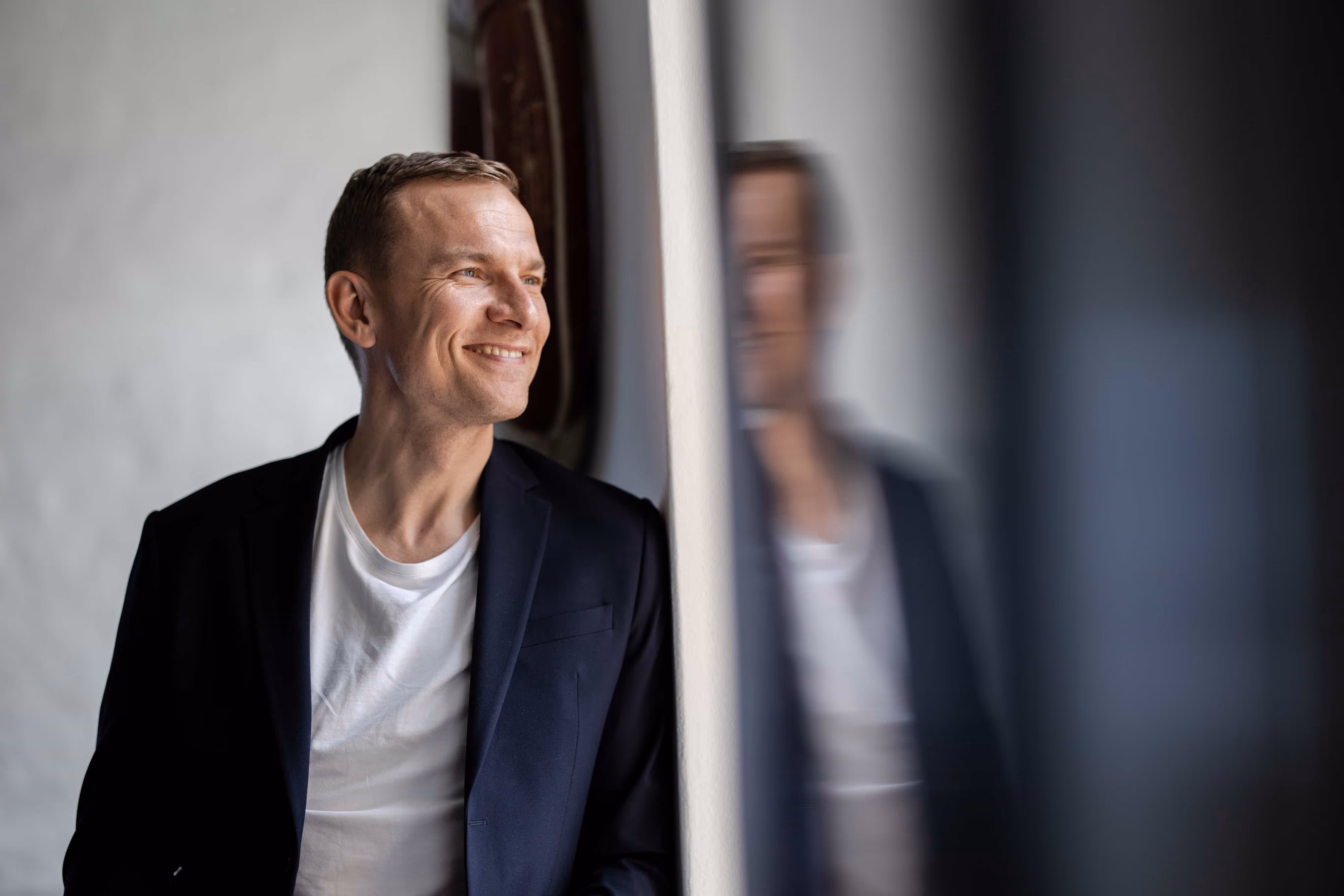 Mand i hvidt T-shirt og mørk blazer smiler og kigger ud ad et vindue med sin refleksion ved siden af.