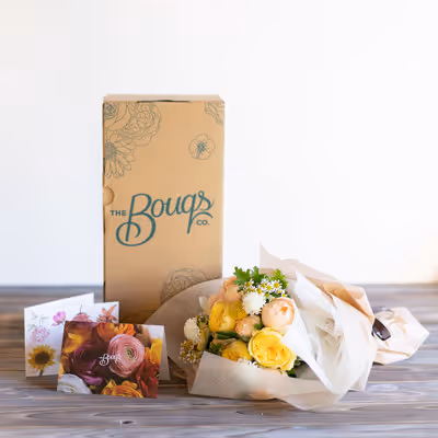 The Bouqs Co. delivery box with a yellow and white bouquet and floral greeting cards on a wooden table.