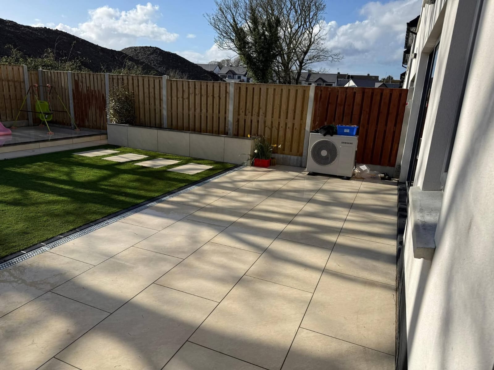 Backyard with beige tile patio, artificial grass section with stepping stones, wooden fence, and a Samsung air conditioning unit.
