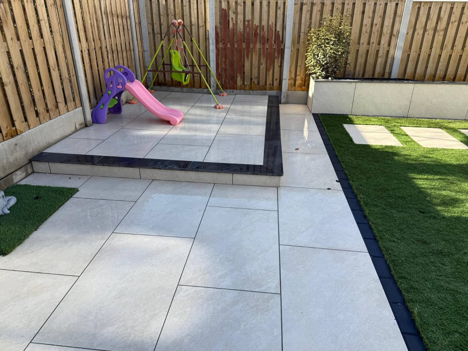 Outdoor patio with beige tiles bordered by dark granite, a raised platform with a pink slide and green swing, and artificial grass with stepping stones.