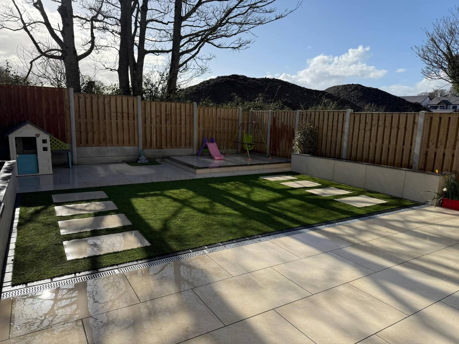 Backyard patio with light stone tiles, an artificial grass lawn with stepping stones, wooden fence, children's play slide and swing, and a small playhouse.