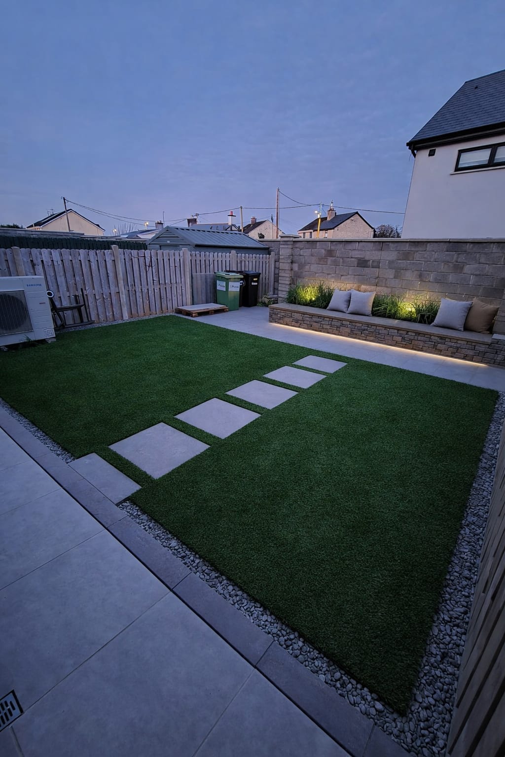 Modern backyard with rectangular artificial grass area, stepping stones, tiled patio, and a stone bench with cushions and LED lighting at dusk.