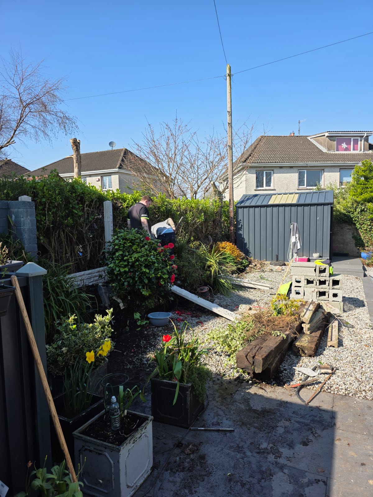 Two people working in a garden with raised beds, surrounded by plants, flowers, gravel, wooden beams, and cinder blocks under a clear blue sky.