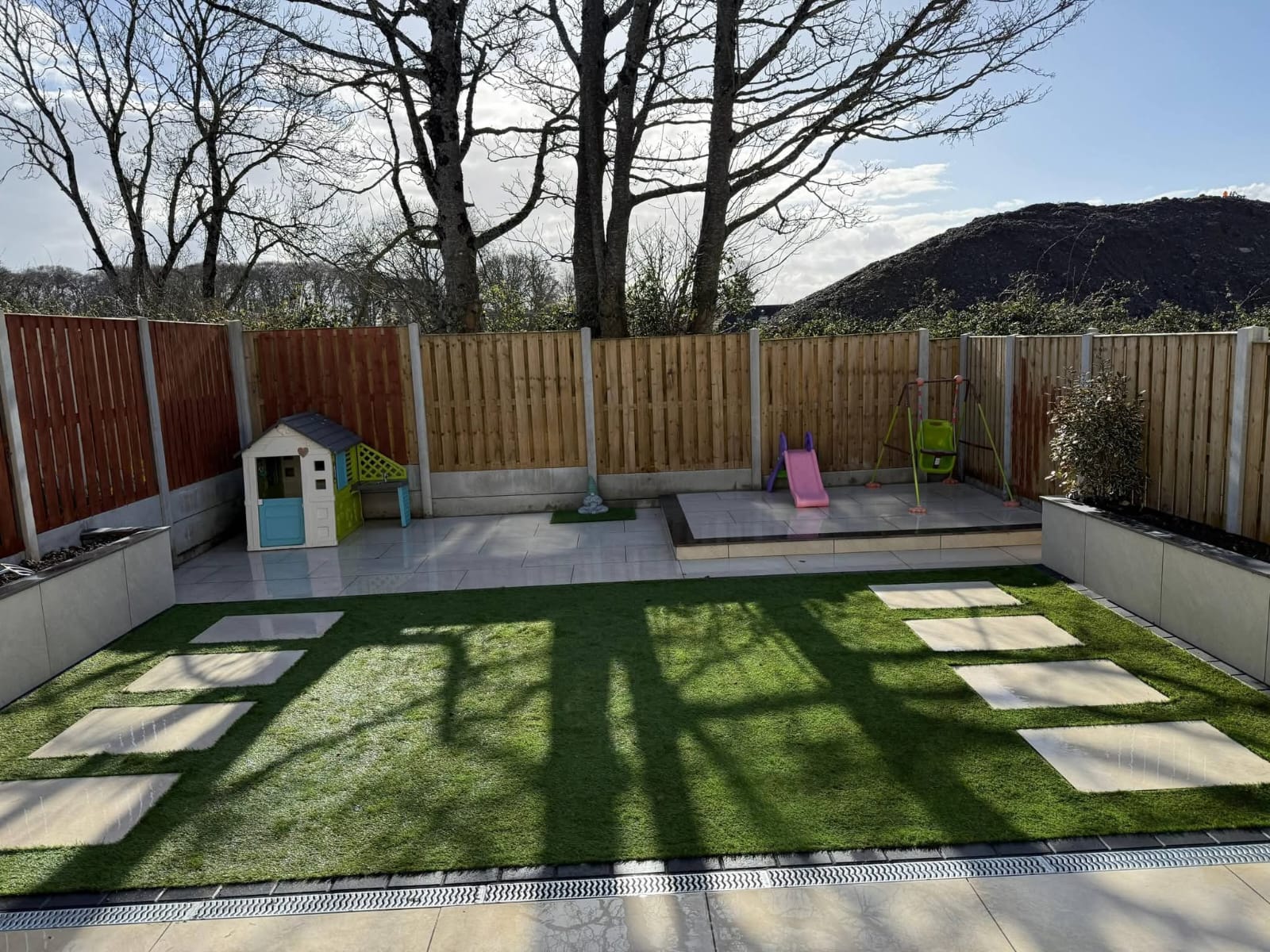 Backyard with artificial grass, stepping stones, fenced edges, a small playhouse, a pink slide, and a green baby swing set on a tiled patio.
