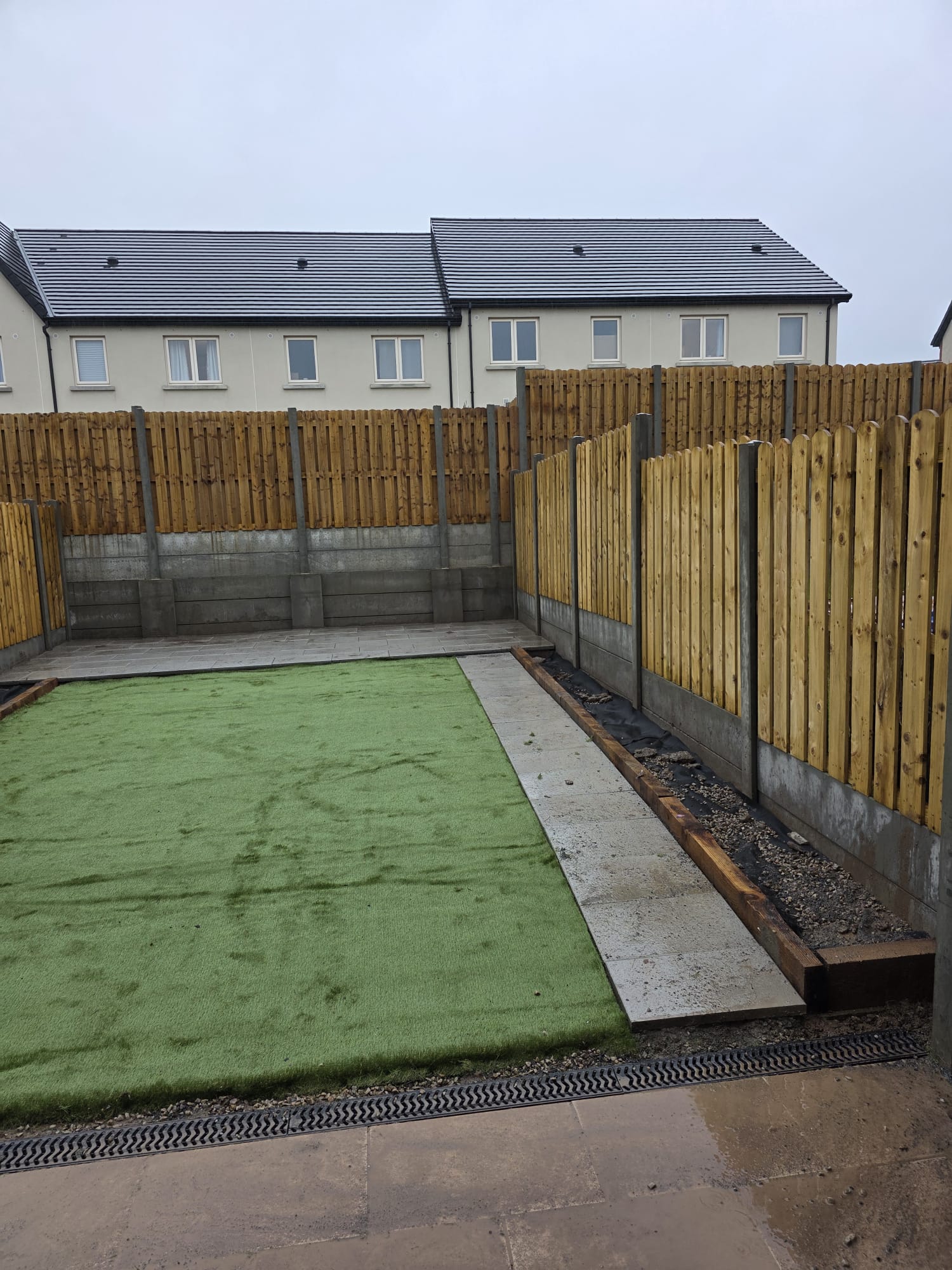 Backyard with green artificial turf, gray paving stones, wooden flower bed borders, and tall wooden fences under an overcast sky.