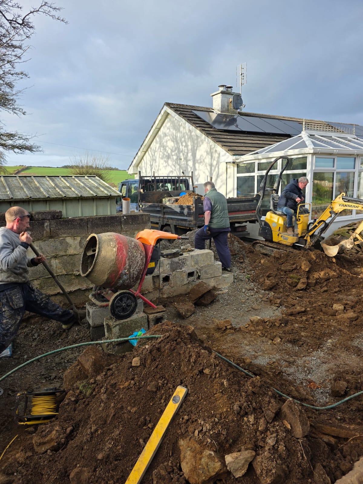 Men working on outdoor construction near a house, with soil piles, a cement mixer, concrete blocks, and a small excavator.