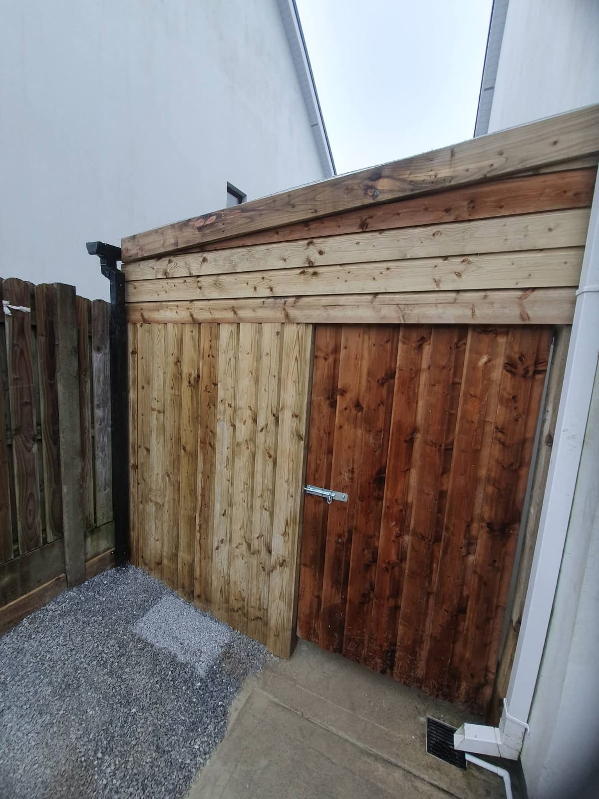 Wooden side gate with a metal latch between two white walls, with gravel and concrete on the ground beside it.