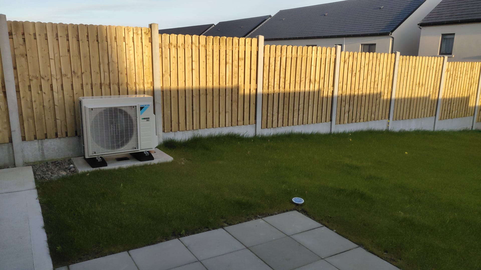 Backyard with a newly installed wooden fence, green grass, concrete patio tiles, and a white Daikin air conditioning unit on a concrete base.