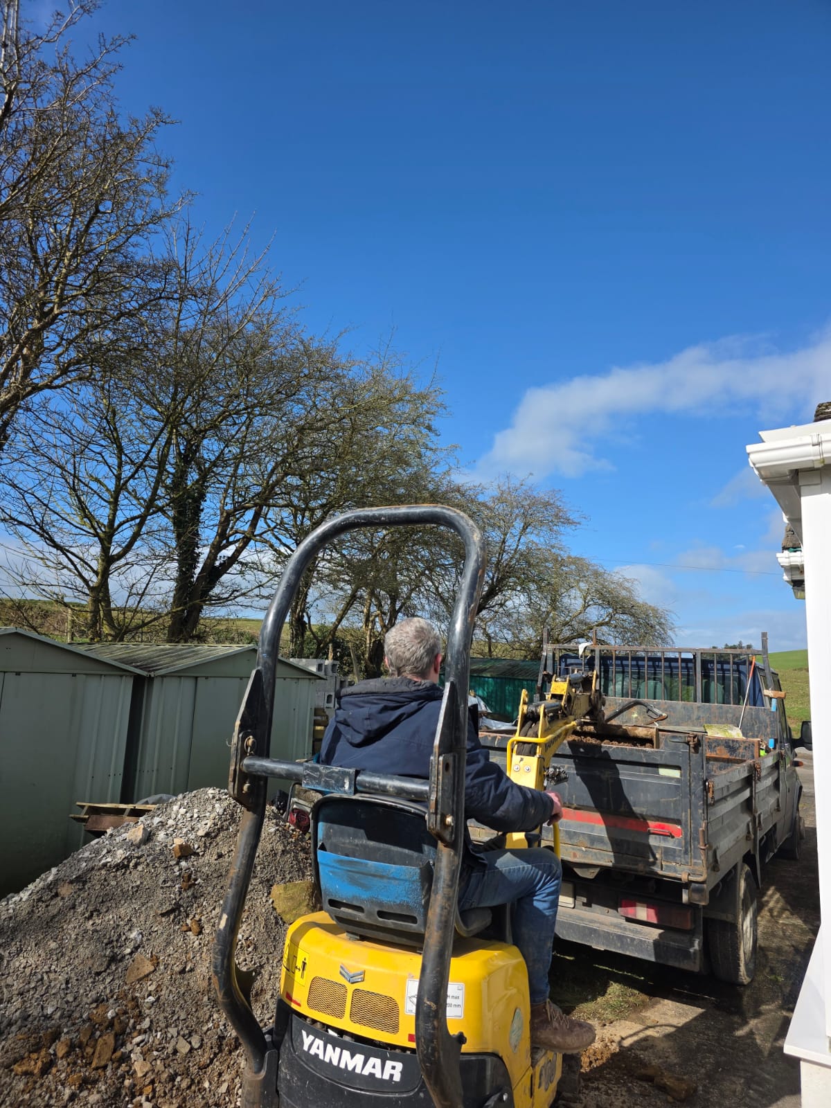Man operating yellow Yanmar mini digger to load gravel into a black truck under a clear blue sky.