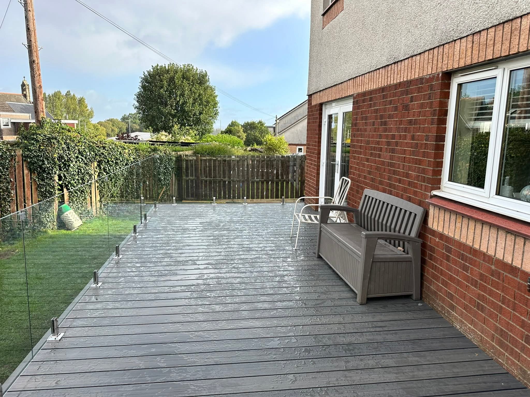 Wet gray composite deck with glass railing, a brown bench, and a white metal chair next to a brick house.