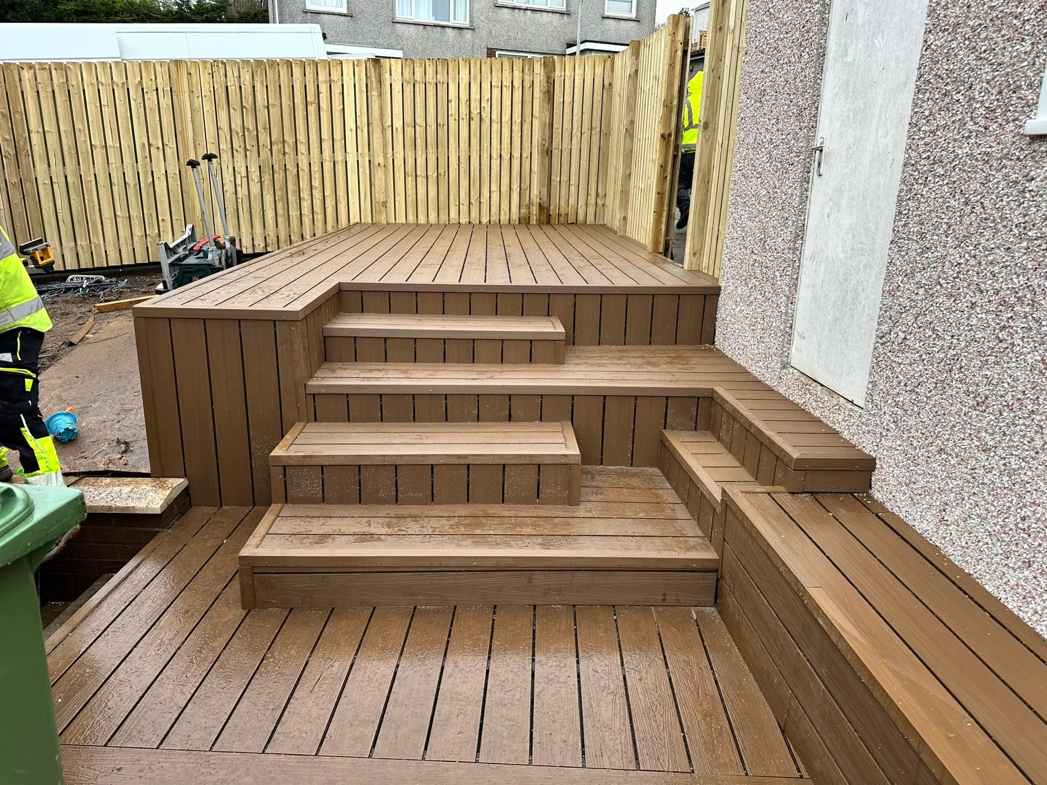 Multi-level brown composite wood deck with stairs and built-in bench against a textured wall and wooden fence background.