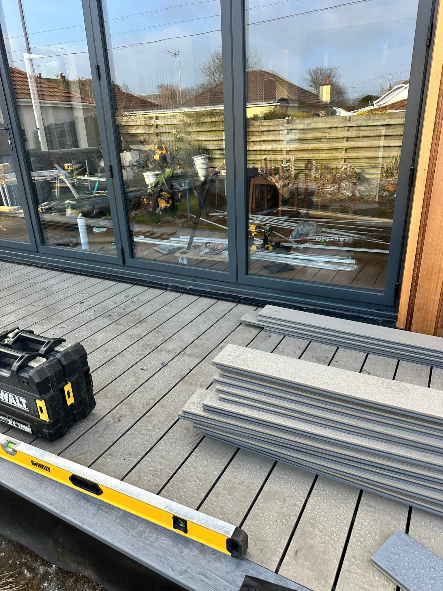 Outdoor deck installation in progress with stacked composite decking boards, a yellow Dewalt level, and a black Dewalt toolbox near glass doors.