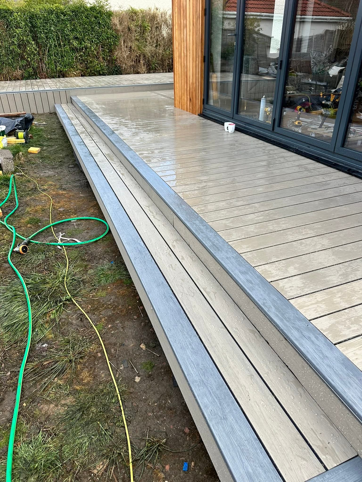 Newly installed wet composite wood deck with steps next to a sliding glass door and garden hose on grass.