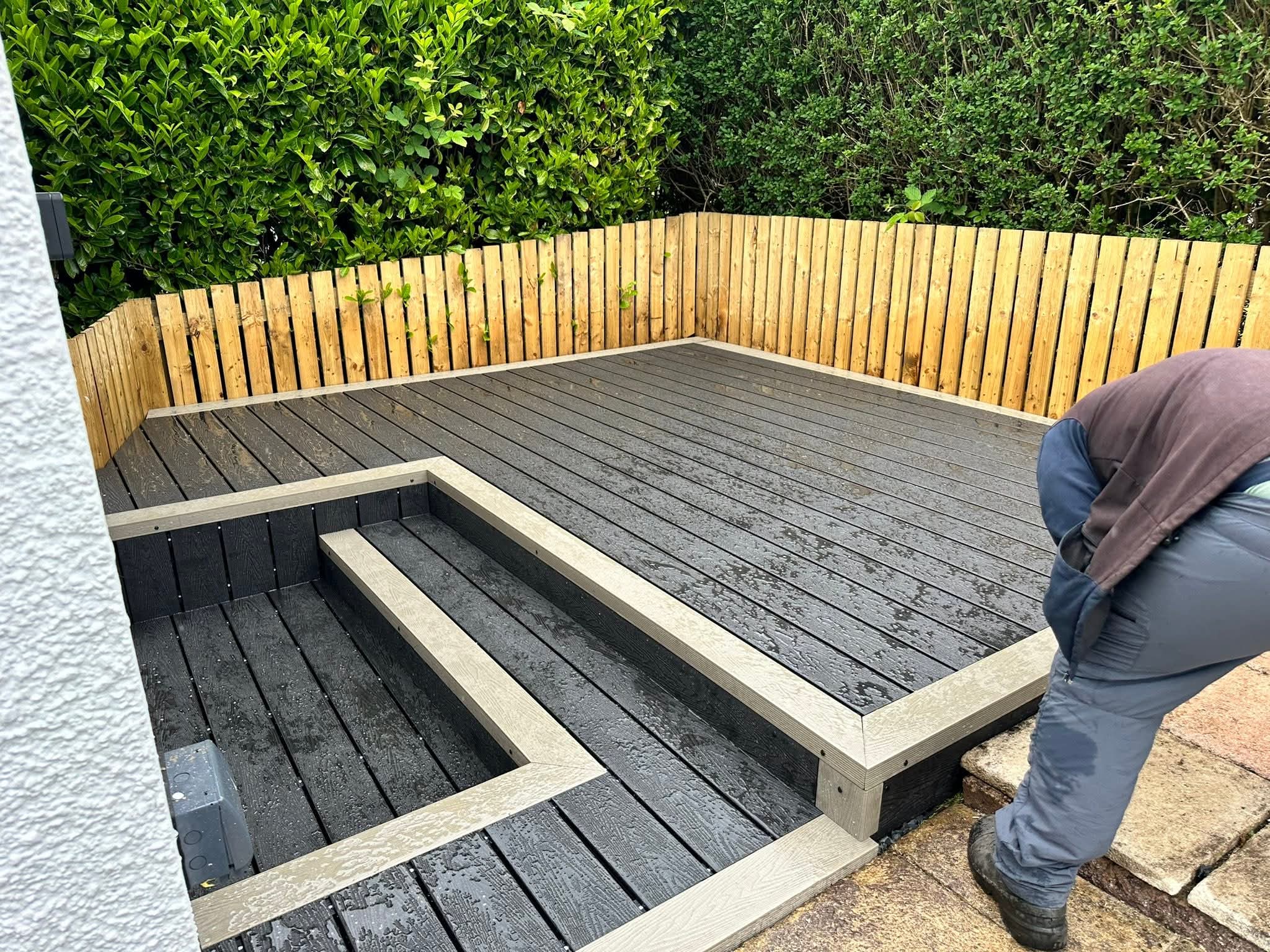 Wet composite wood deck with two beige-bordered steps and a surrounding wooden fence with green bushes behind it.