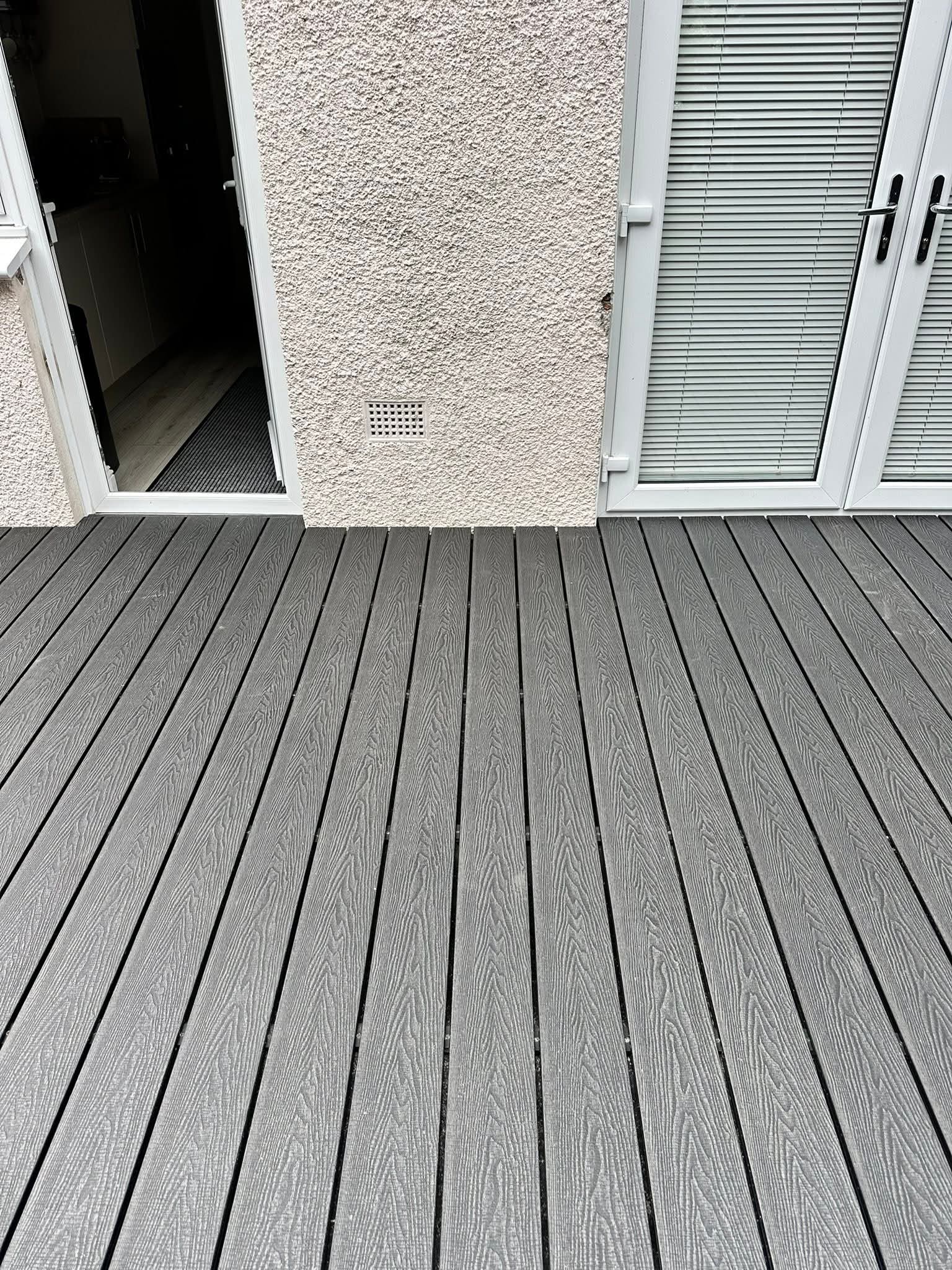 Gray composite decking in front of a white textured wall with an open door on the left and closed glass doors with blinds on the right.