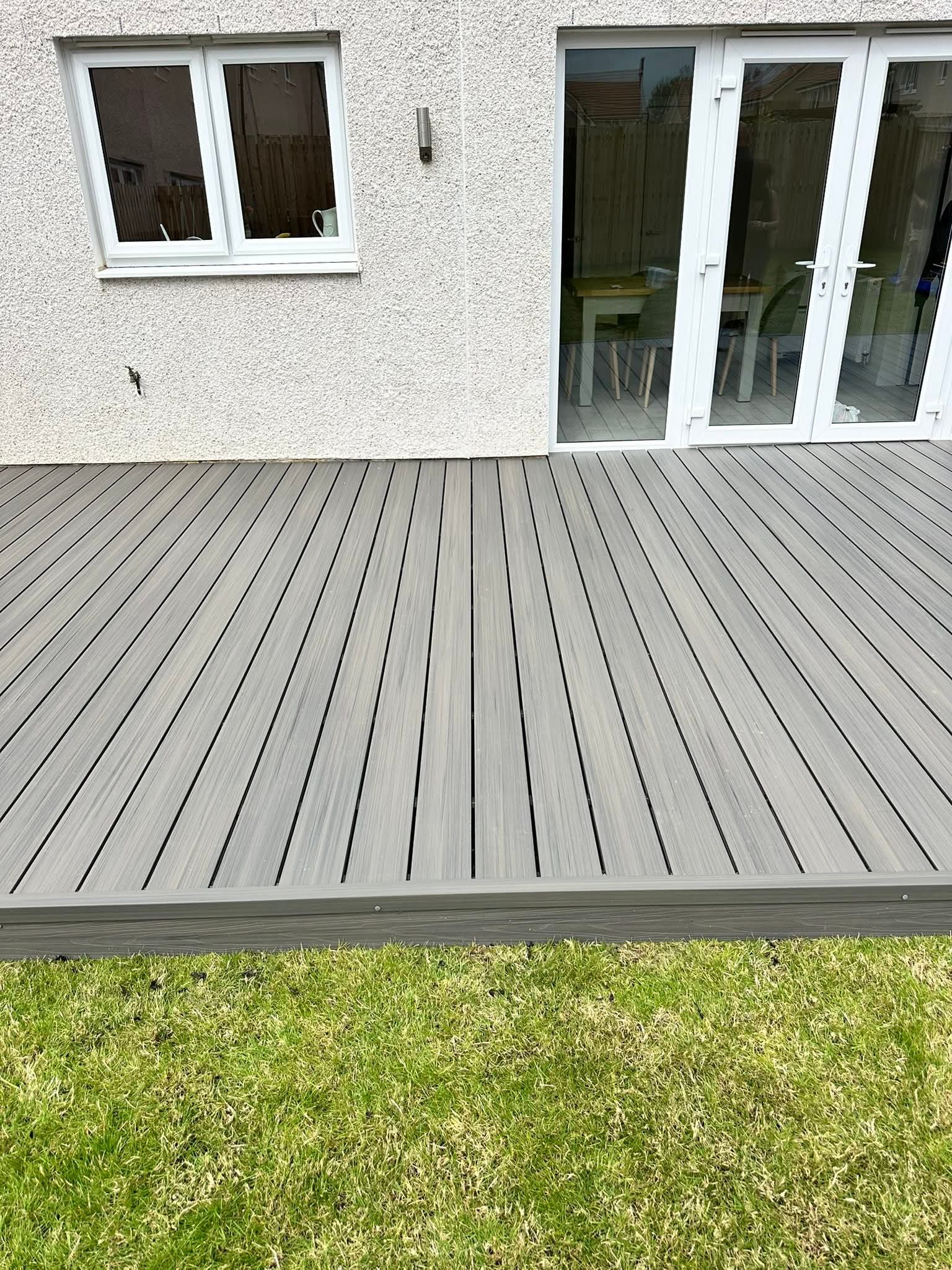 Gray composite decking in a backyard next to a white wall with windows and glass doors, and green grass in the foreground.