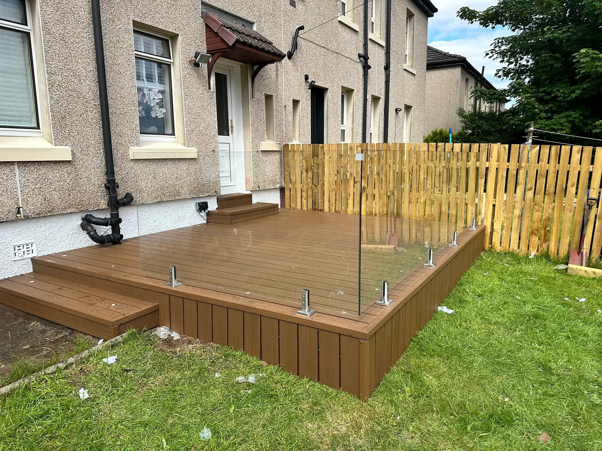 Brown composite deck with glass railing in a backyard next to a beige house and green lawn.