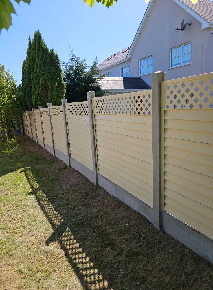Newly installed wooden fence panel with lattice top leaning against a dark wooden fence on a paved patio with a coiled green garden hose and measuring tape nearby.