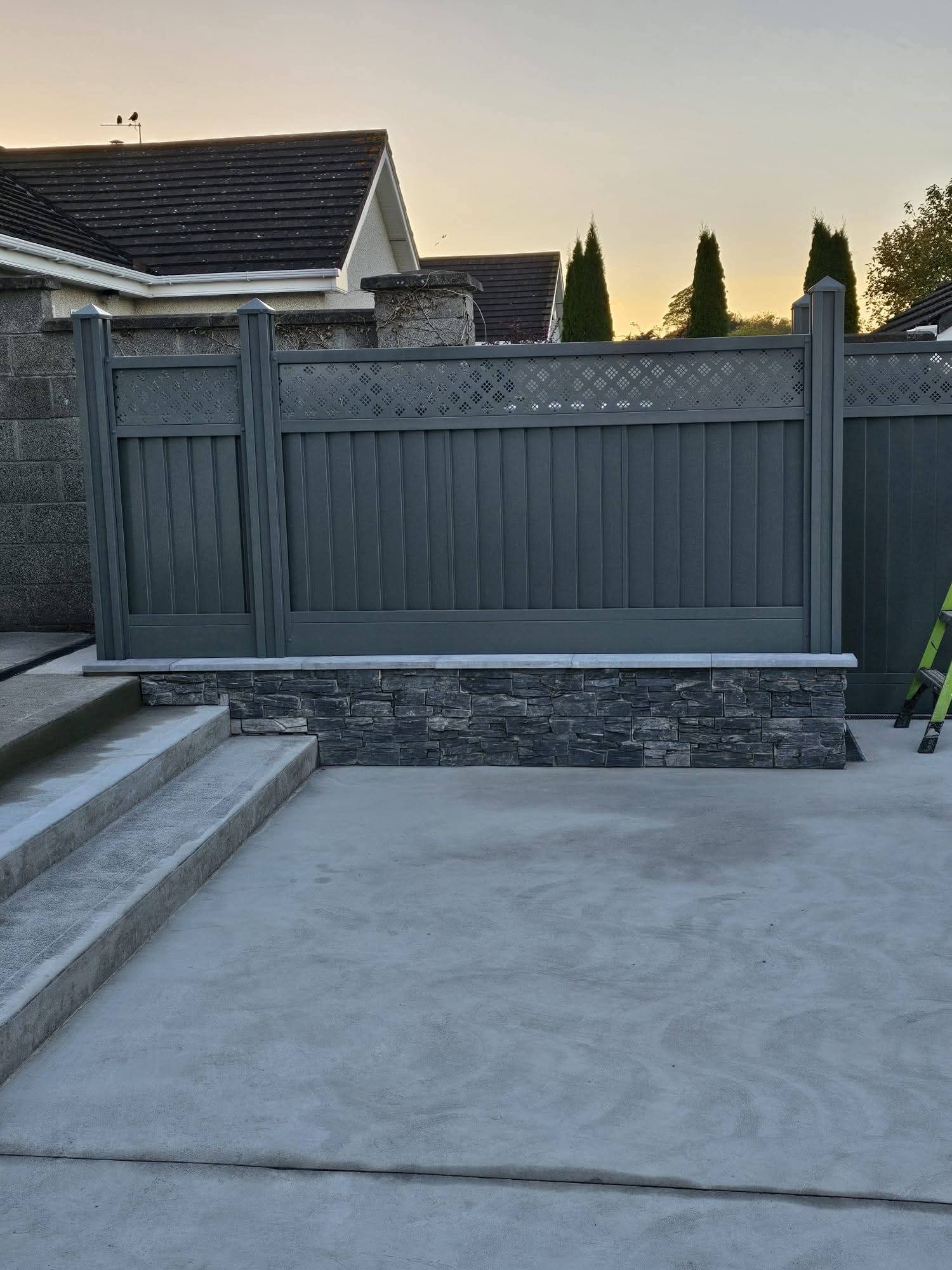 Light-colored wooden garden fence panel with lattice top placed on a stone patio next to a coiled green garden hose and a measuring tape.