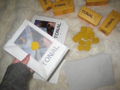 Hand holding a translucent envelope sealed with a yellow wax stamp over a toner booklet, with more envelopes, yellow wax seals, and TONAL Sunveil product boxes on a light fuzzy surface.