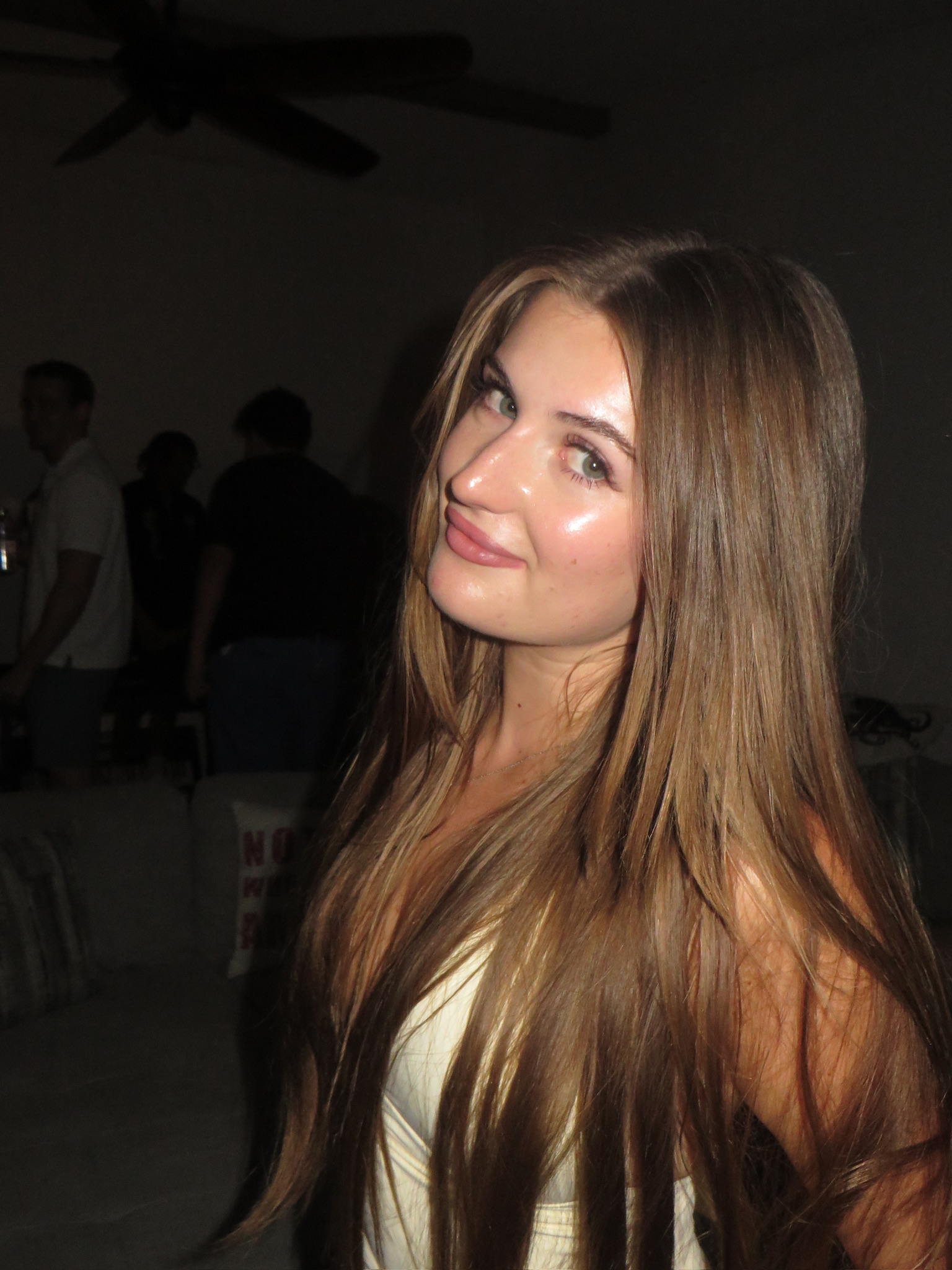 Young woman with long brown hair smiling and tilting her head in a dimly lit room with people in the background.