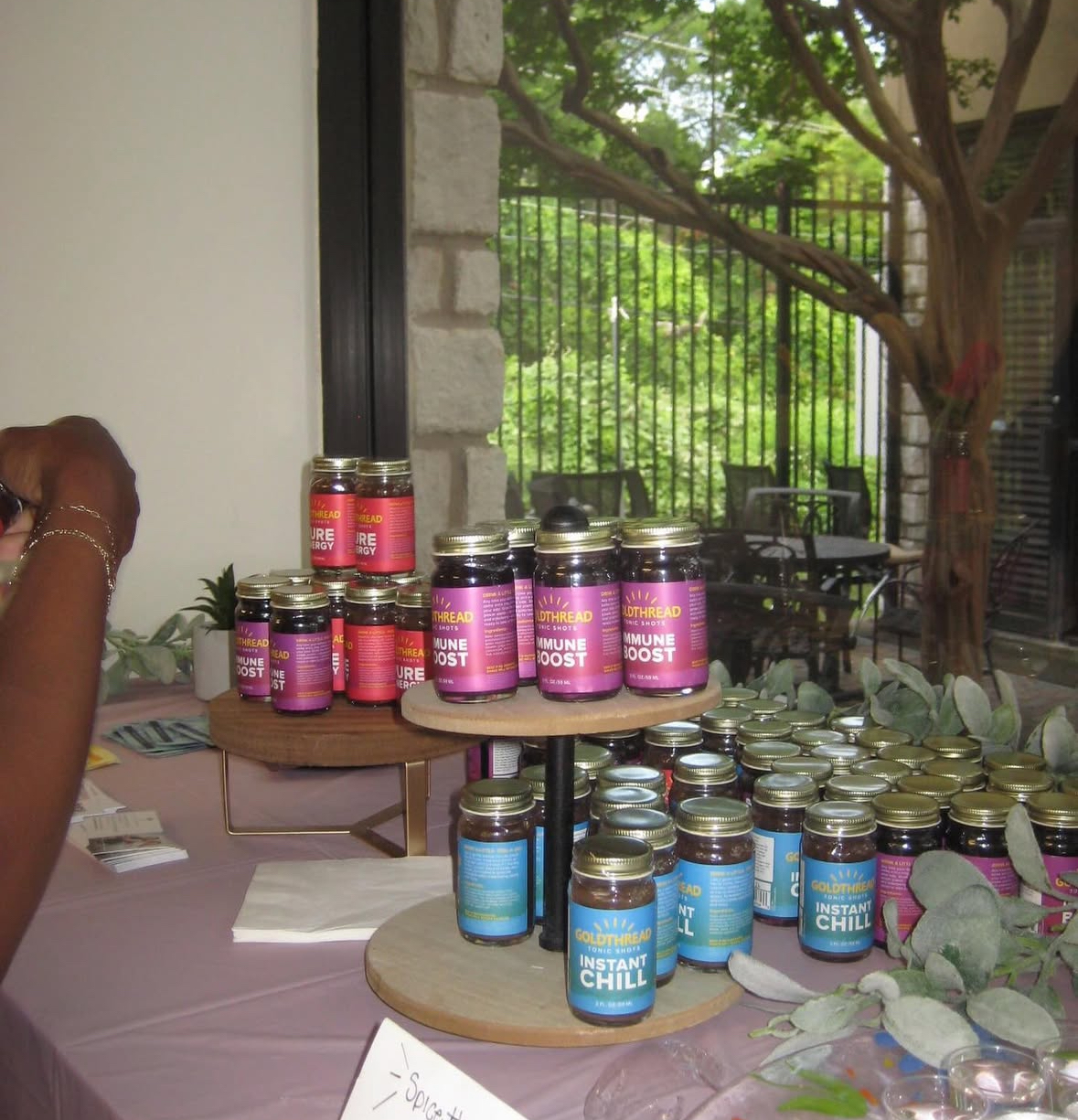 Display of small bottles labeled 'Immune Boost' and 'Instant Chill' tonic shots on wooden tiered stands at an indoor event.