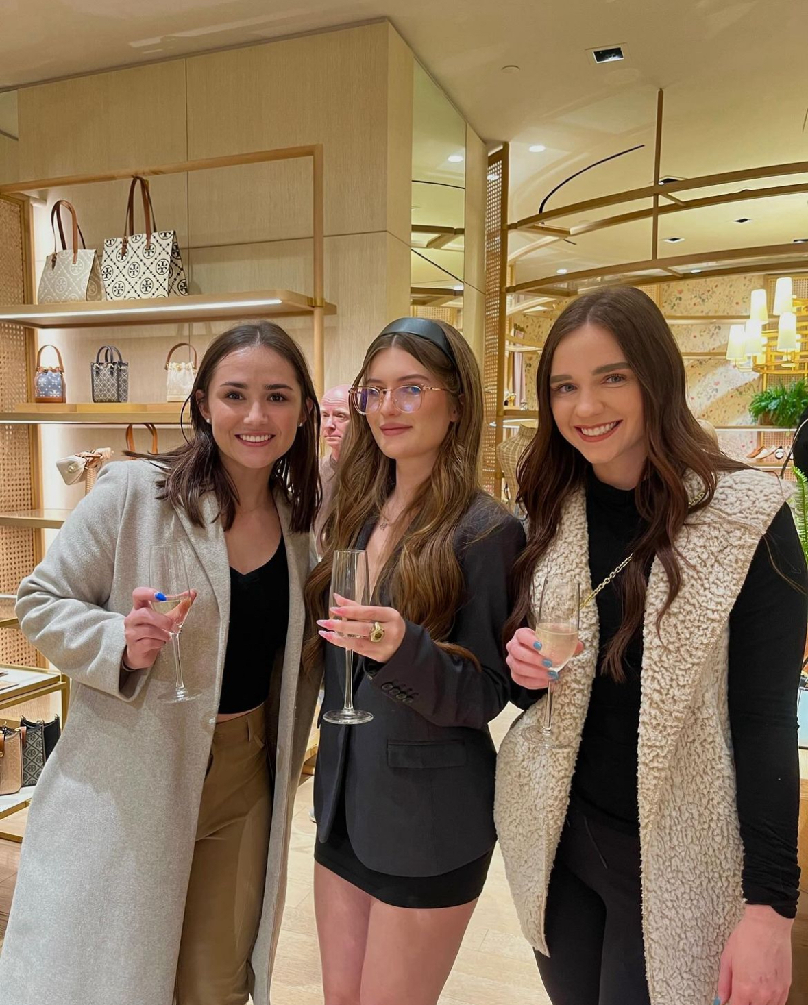 Three young women smiling and holding champagne flutes inside a stylish boutique with handbags displayed in the background.