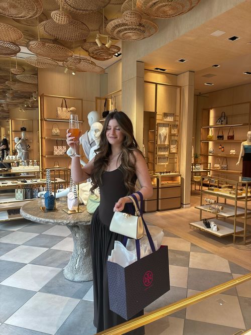 Woman in black dress holding a white handbag and a shopping bag in a luxury boutique, raising a glass with an orange drink.