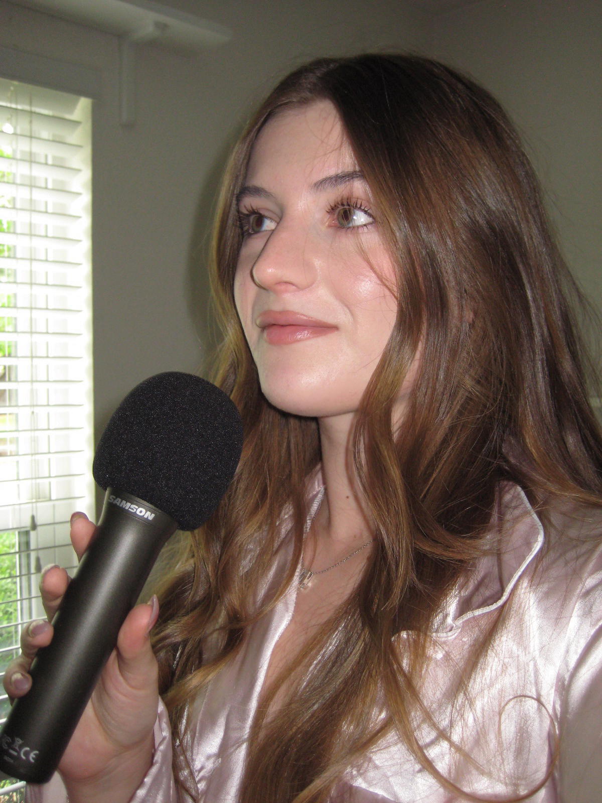 Young woman with long brown hair holding a black Samson microphone near her mouth indoors by a window with blinds.