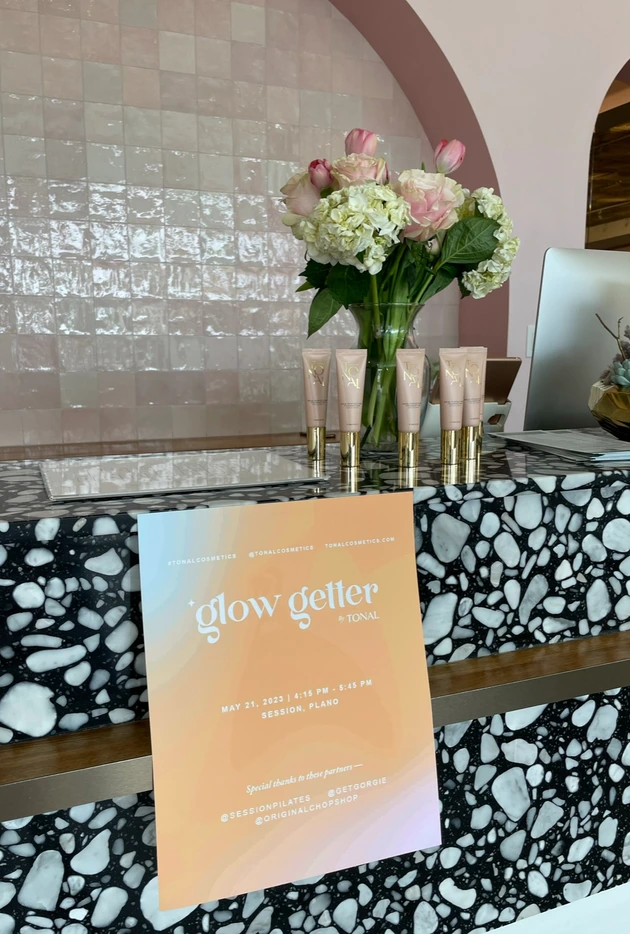 Marble-patterned counter with a vase of pink and white flowers, tubes of cosmetics, and a sign that reads 'glow getter by TONAL' with event details.