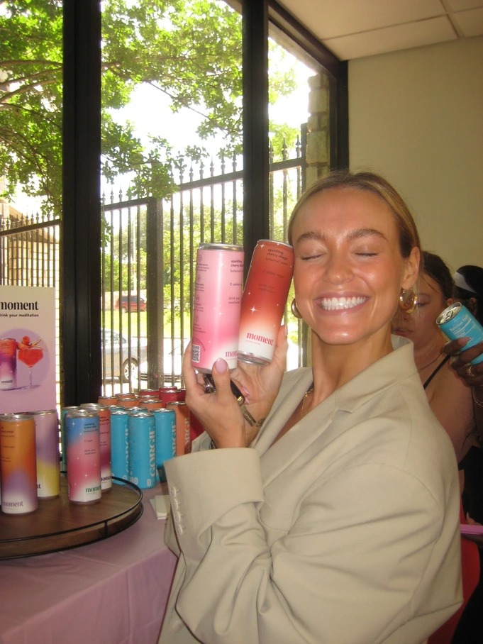 Smiling woman in beige jacket holding two cans of Moment sparkling cherry seltzer near a display of colorful canned drinks.