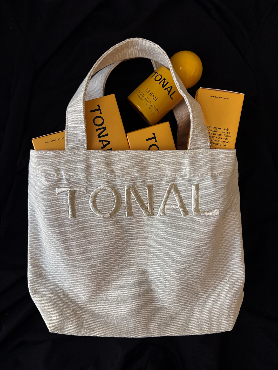 Beige canvas tote bag with TONAL embossed on it, filled with yellow TONAL cosmetics packaging and a round yellow bottle.