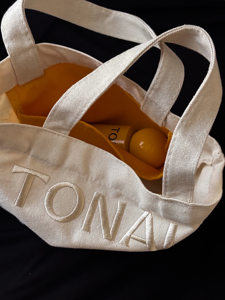 Close-up of an off-white canvas tote bag with wide straps and the text 'TONAI' embroidered on it, containing a yellow item inside.