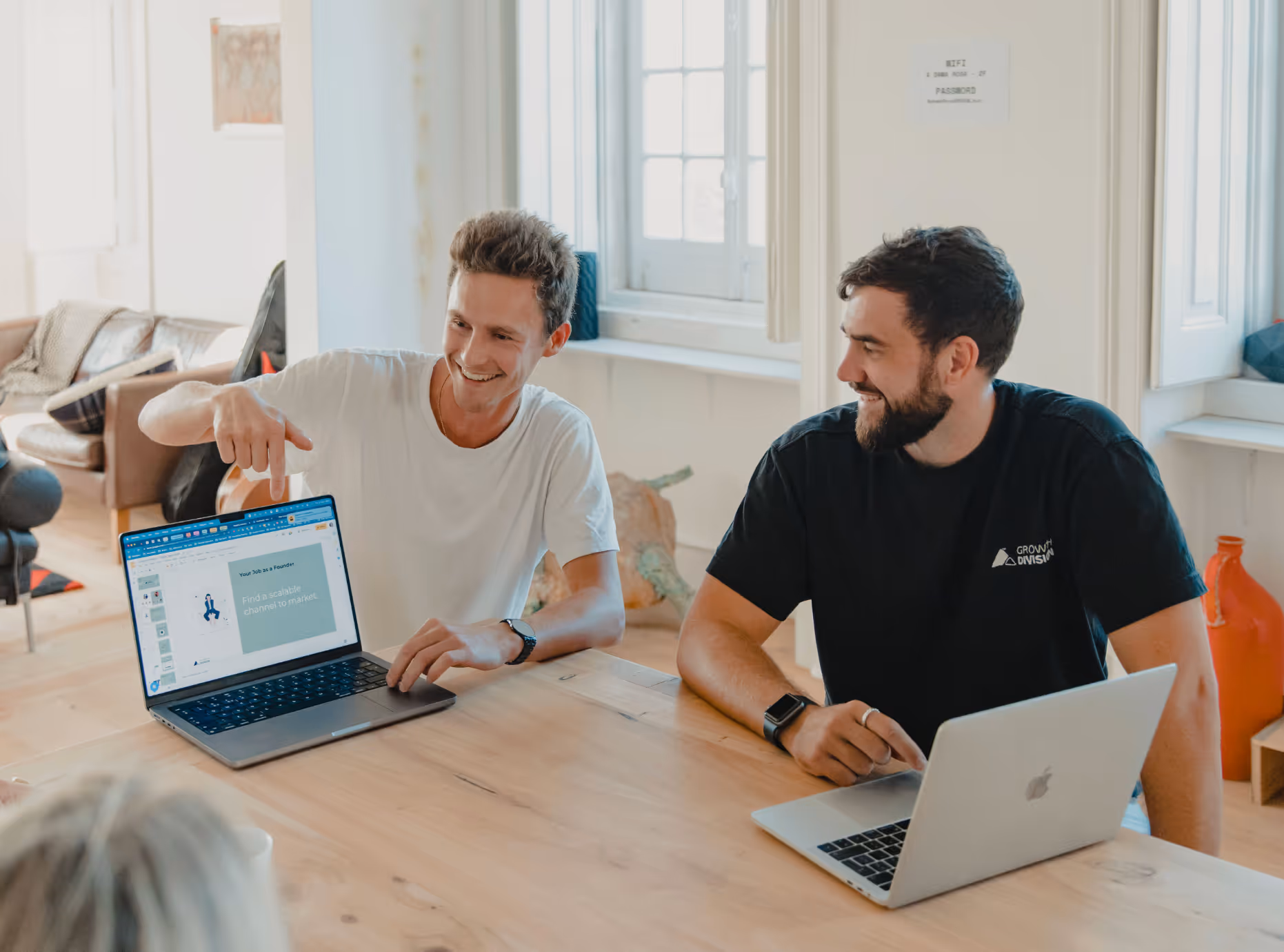 Two men sitting at a wooden table with laptops, one pointing at a presentation slide on his laptop screen that reads 'Find a scalable channel to market.'