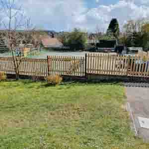 Low palisade fence enclosing a village green with houses in the background.
