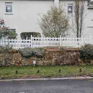 White palisade fence bordering a driveway with shrubs and lawn.