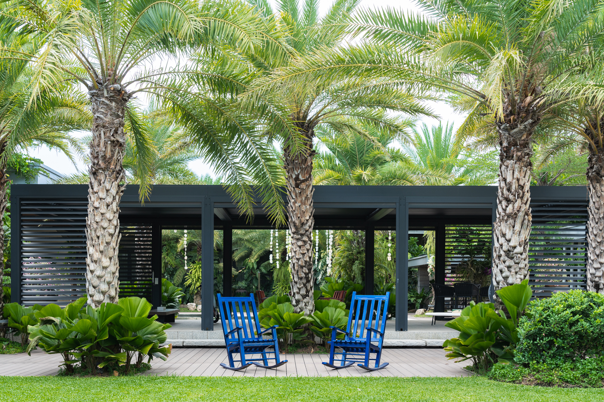 Lexus House — In front of the outdoor pavilion are two blue rocking chairs that sit on a wooden deck, surrounded by palm trees, providing a comfortable spot to relax and take in the natural surroundings.