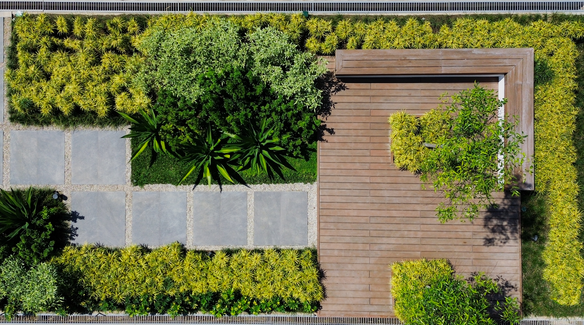 Aerial view of the complex' linear garden features concrete squares on a gravel bed leading to a wooden deck with benches and tree pits, framed by fine-textured shrubs.