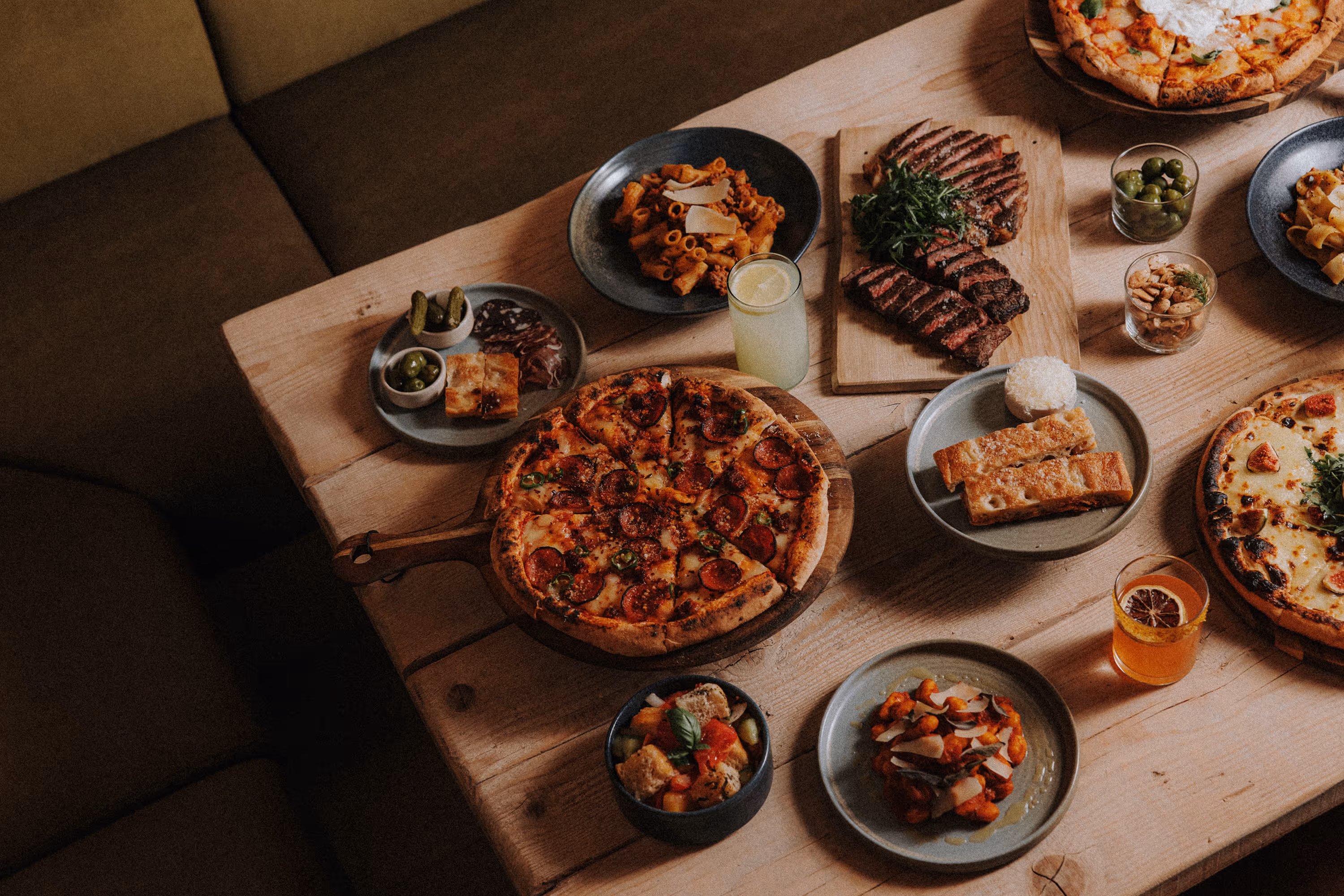Wood table with pepperoni pizza, steak, pasta, breadsticks, olives, salad, drinks, and appetizers.