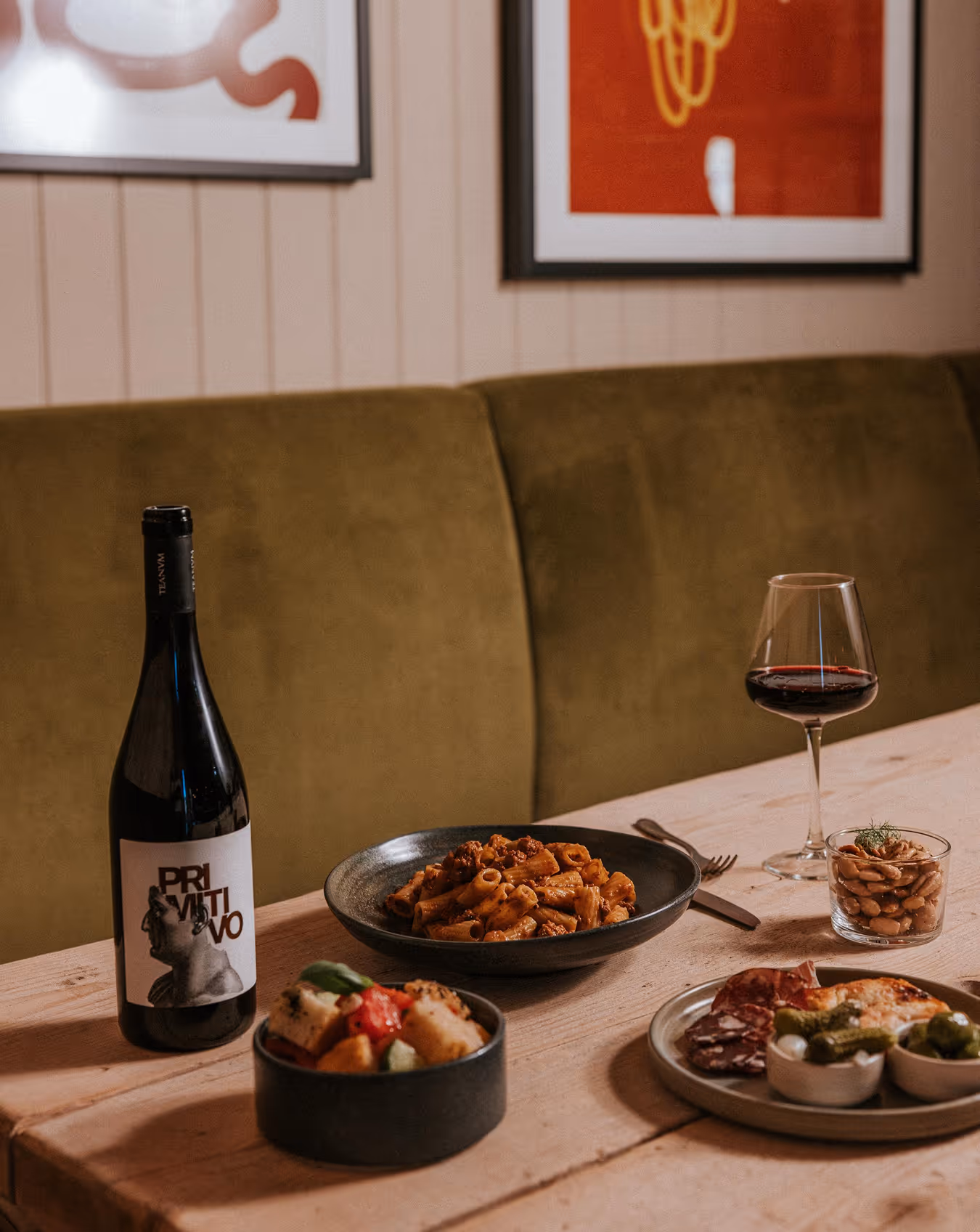 Table with a bottle of red wine, a glass of wine, a pasta dish, bread salad, and charcuterie plate.