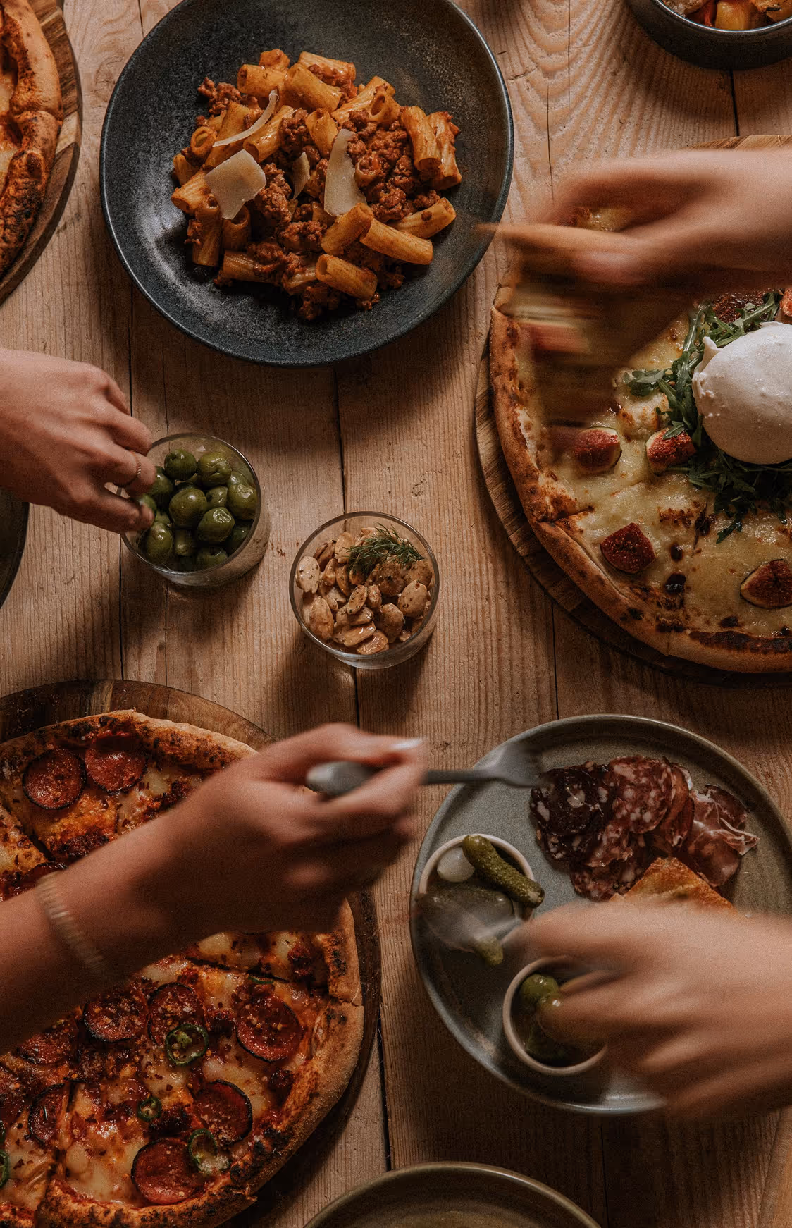Hands reaching for plates with pepperoni pizza, pasta, olives, nuts, and charcuterie on wooden table.
