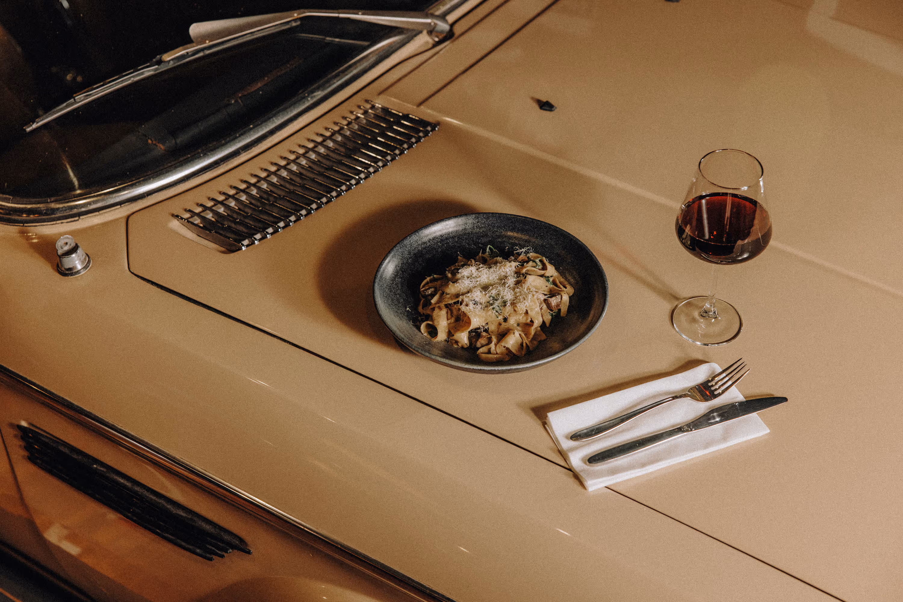 Black plate of pasta with grated cheese and mushrooms on a beige car hood with wine glass and cutlery.
