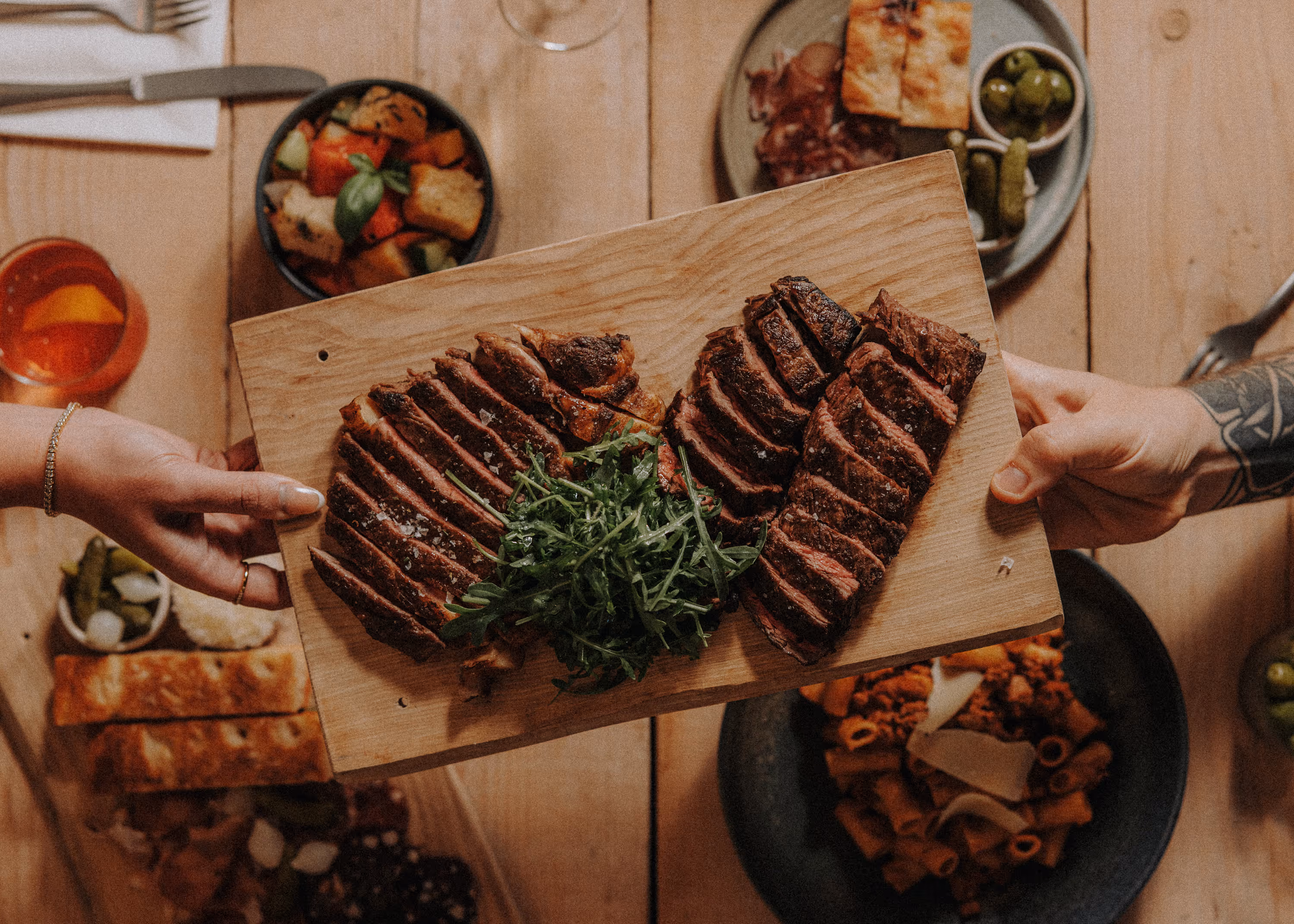 Two hands holding a wooden board with sliced grilled steaks and a pile of greens in the center.