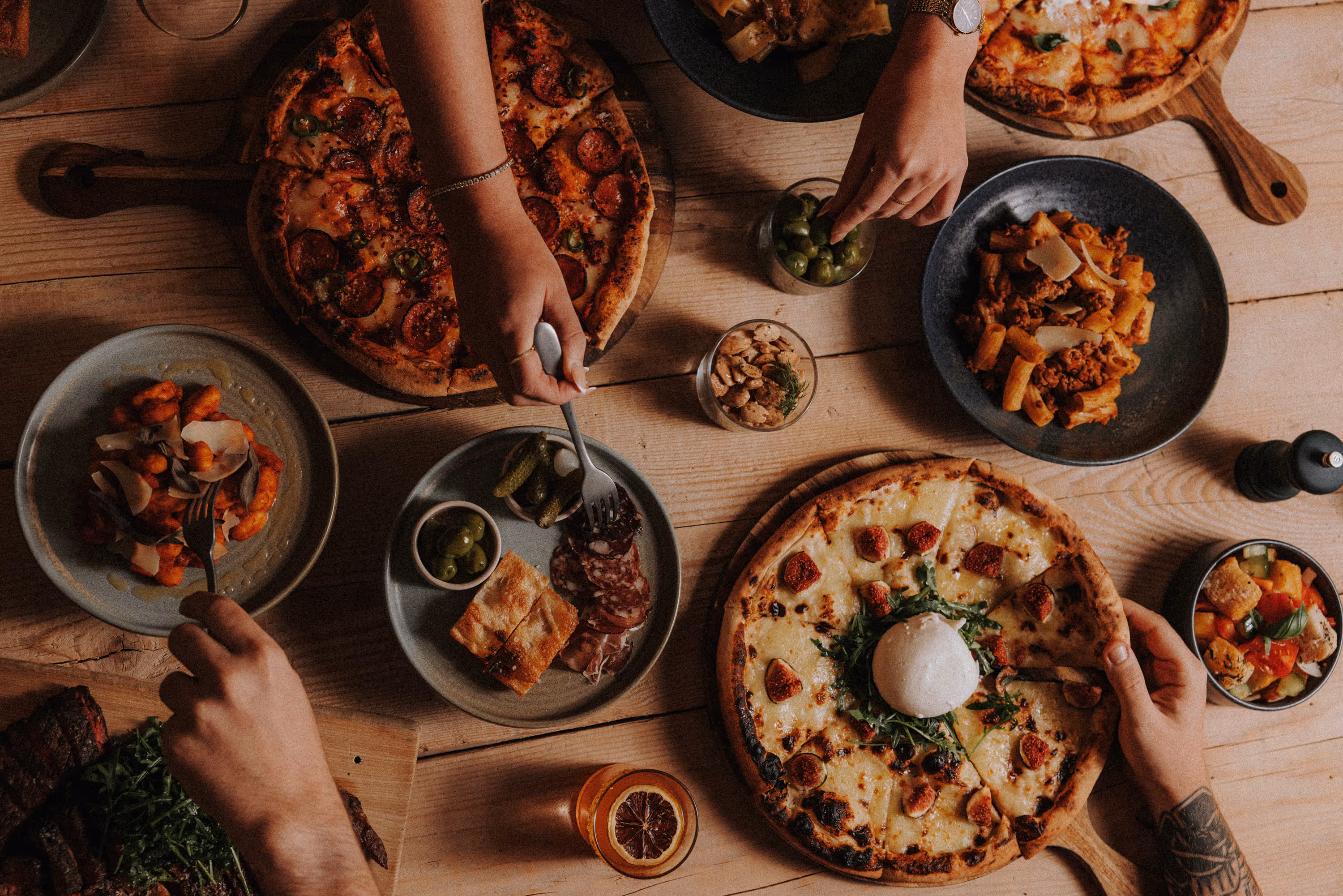 Hands reaching for slices of pepperoni and cheese pizzas on a wooden table with various dishes.