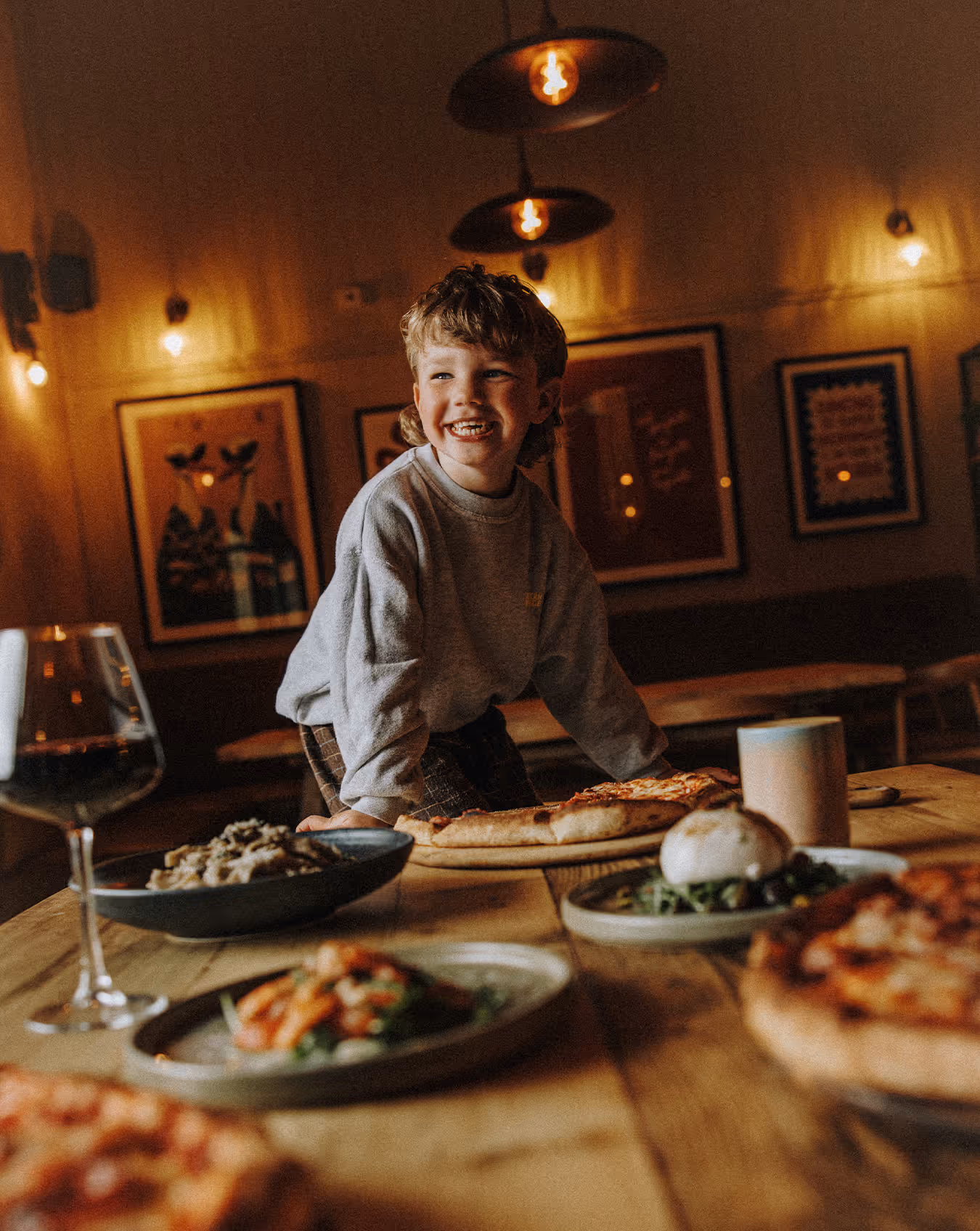 Albert leaning on table with plates of pizza, salad, and a glass of red wine in warm light.