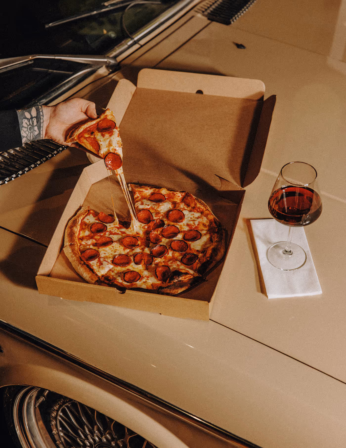 Hand lifting a slice of pepperoni pizza from a box on a car hood next to a glass of red wine.