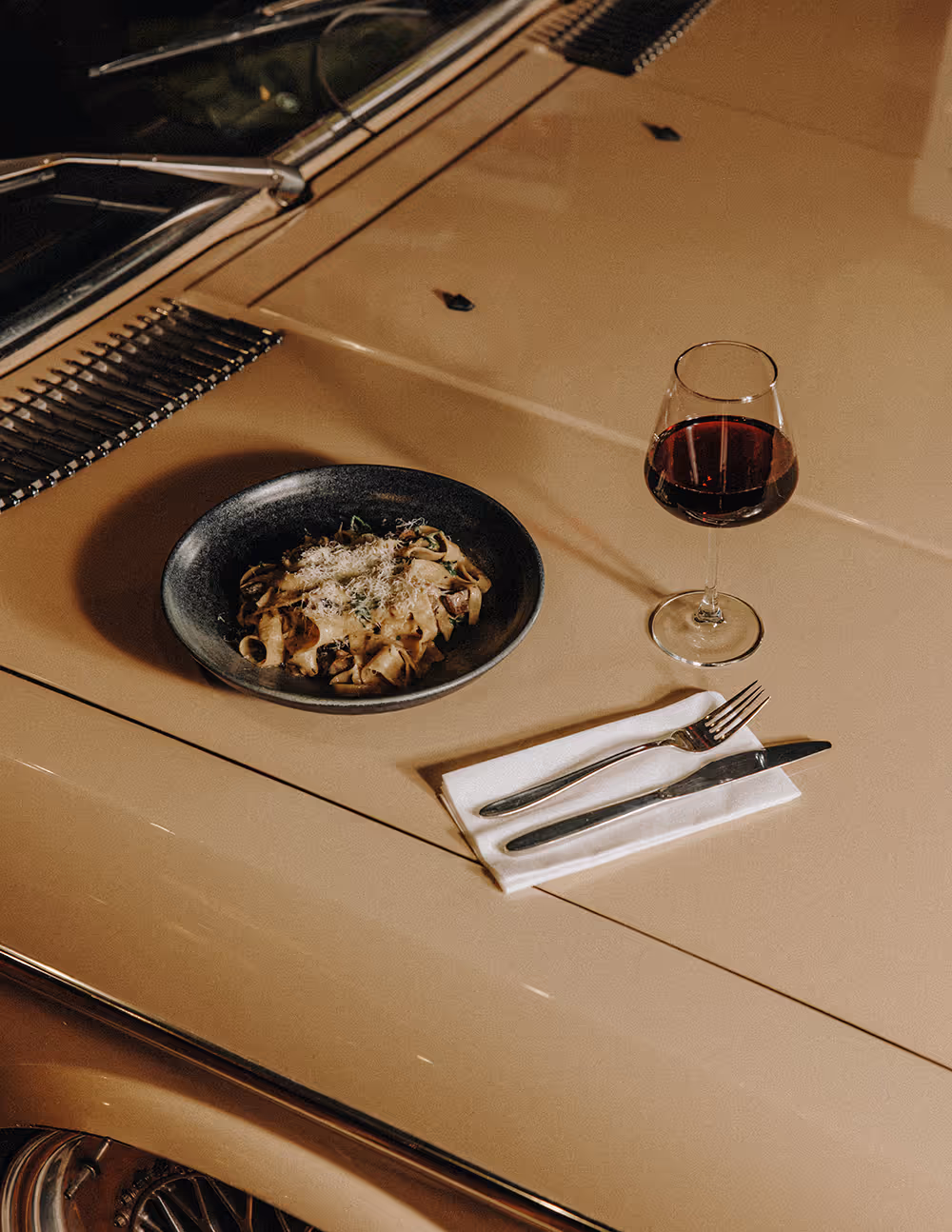 Plate of pasta with grated cheese, glass of red wine, and silverware on napkin on car hood.