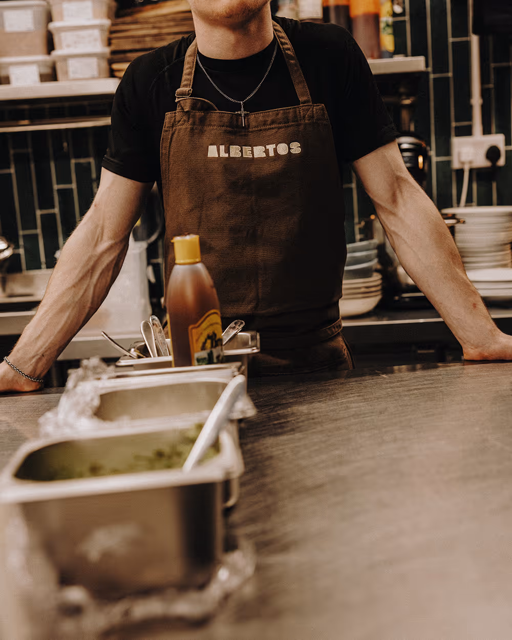Person wearing a brown Alberto's apron stands behind a counter with utensils and condiment bottles.