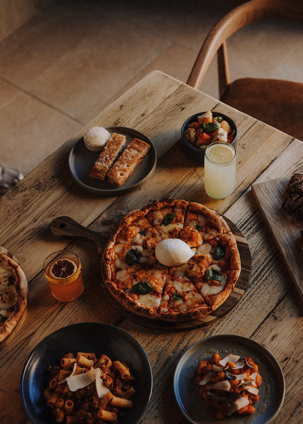 Wood-fired pizza with mozzarella, pasta dishes, breadsticks, salad, and drinks on a wooden table.