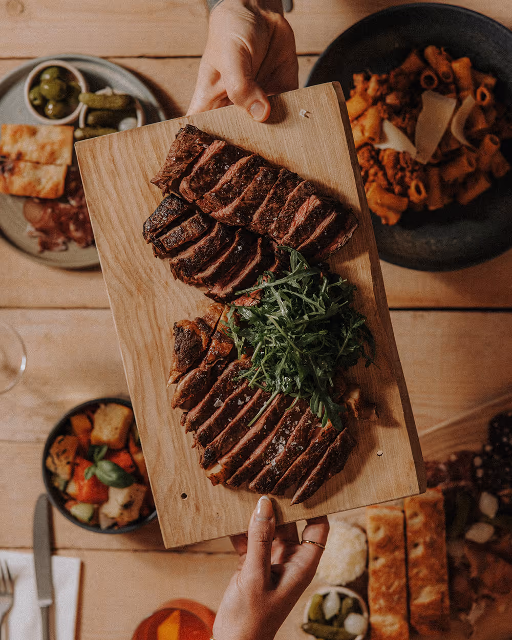 Two hands hold a wooden board with sliced grilled steak and a pile of fresh greens.